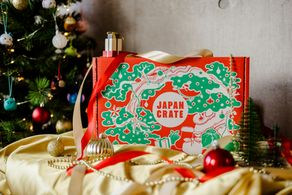 The Christmas Gift Box from Japan Crate contains a variety of Japanese snacks and is placed on a table alongside a Christmas tree.